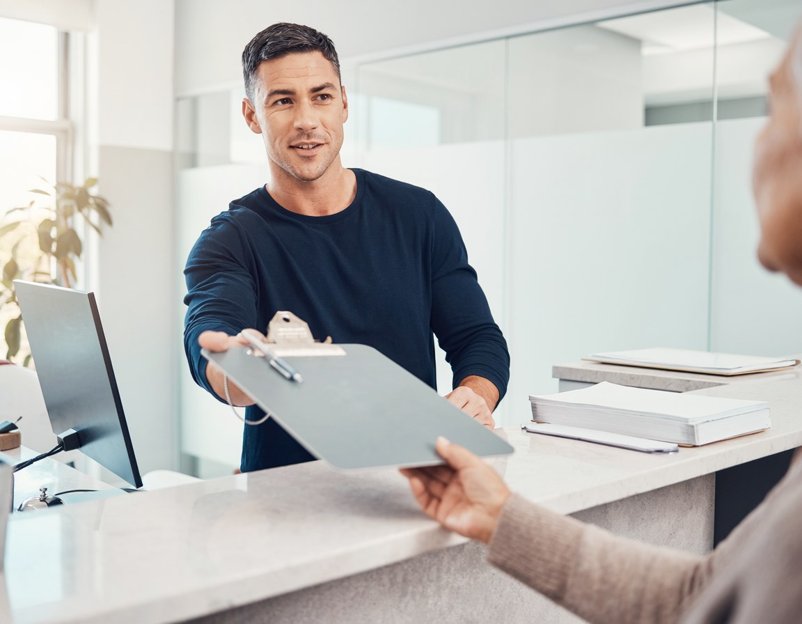 A front desk staff member handing a clipboard across the counter to a patient